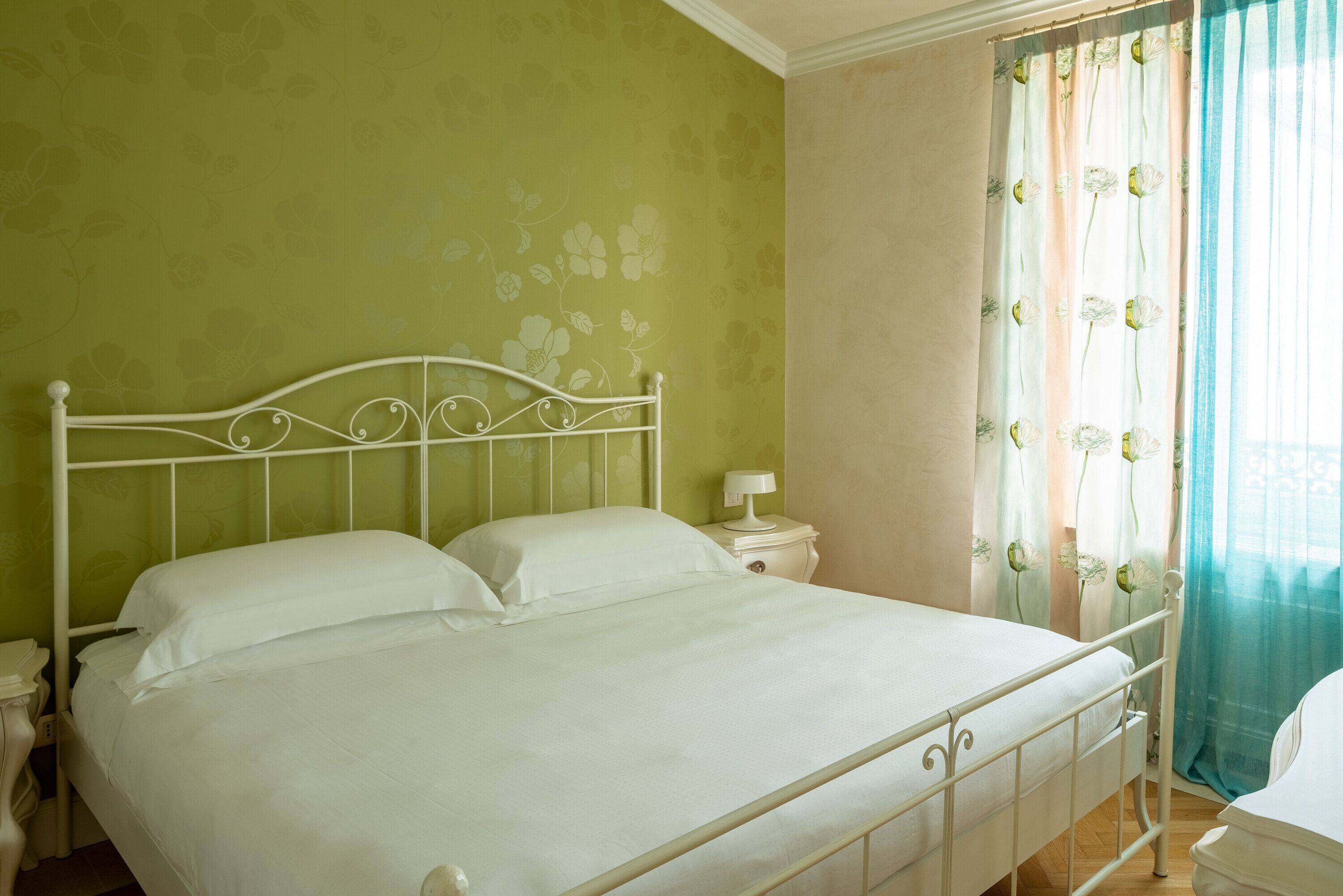 Una cama con estructura metálica, ropa de cama blanca, una mesita de noche con lámpara y una ventana con cortinas florales.