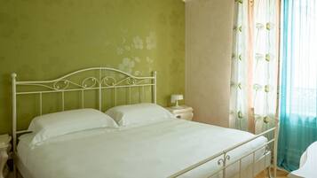 Una cama con estructura metálica, ropa de cama blanca, una mesita de noche con lámpara y una ventana con cortinas florales.