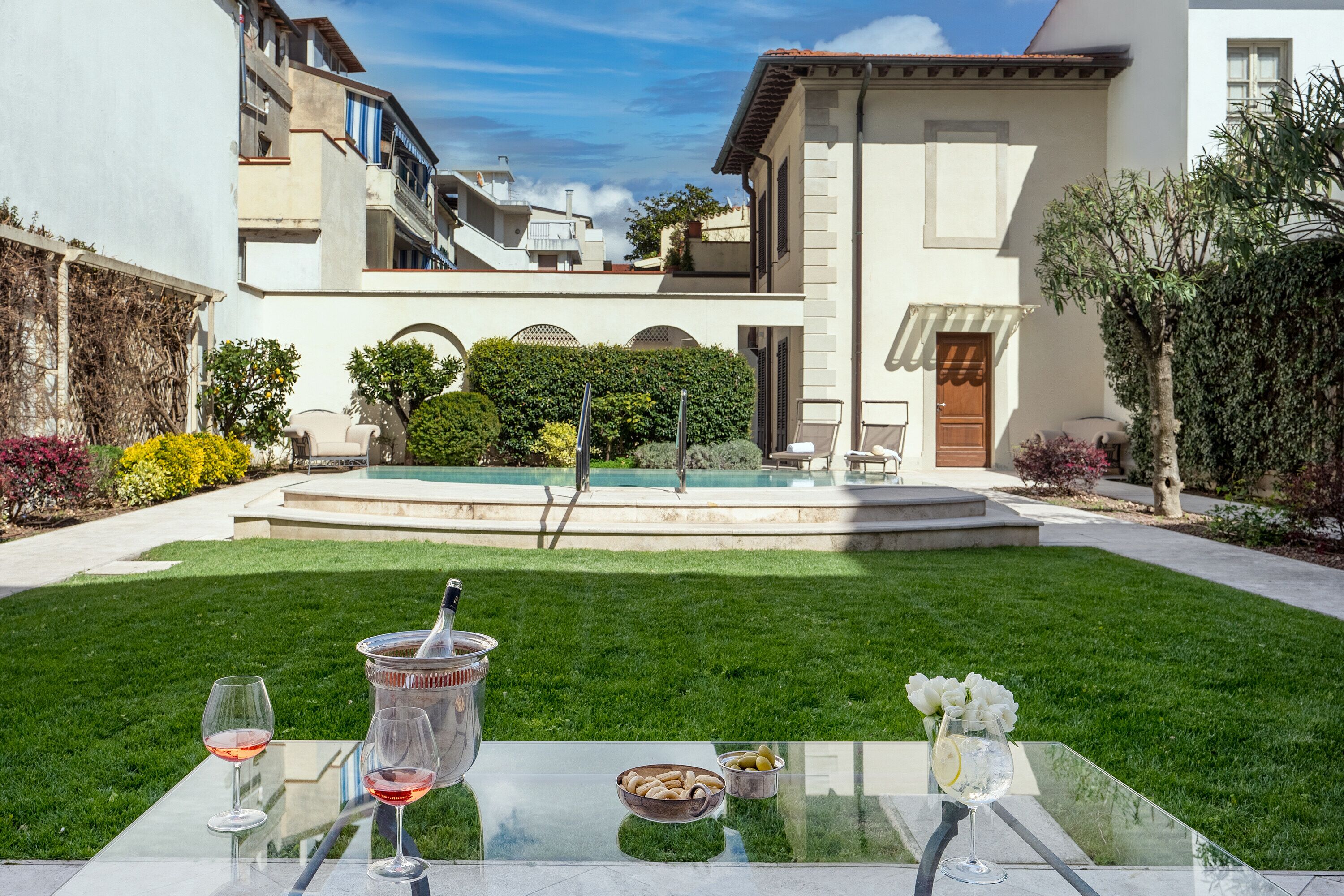 A glass table with a bottle, glasses, and a bowl of snacks in a courtyard with a pool and greenery.