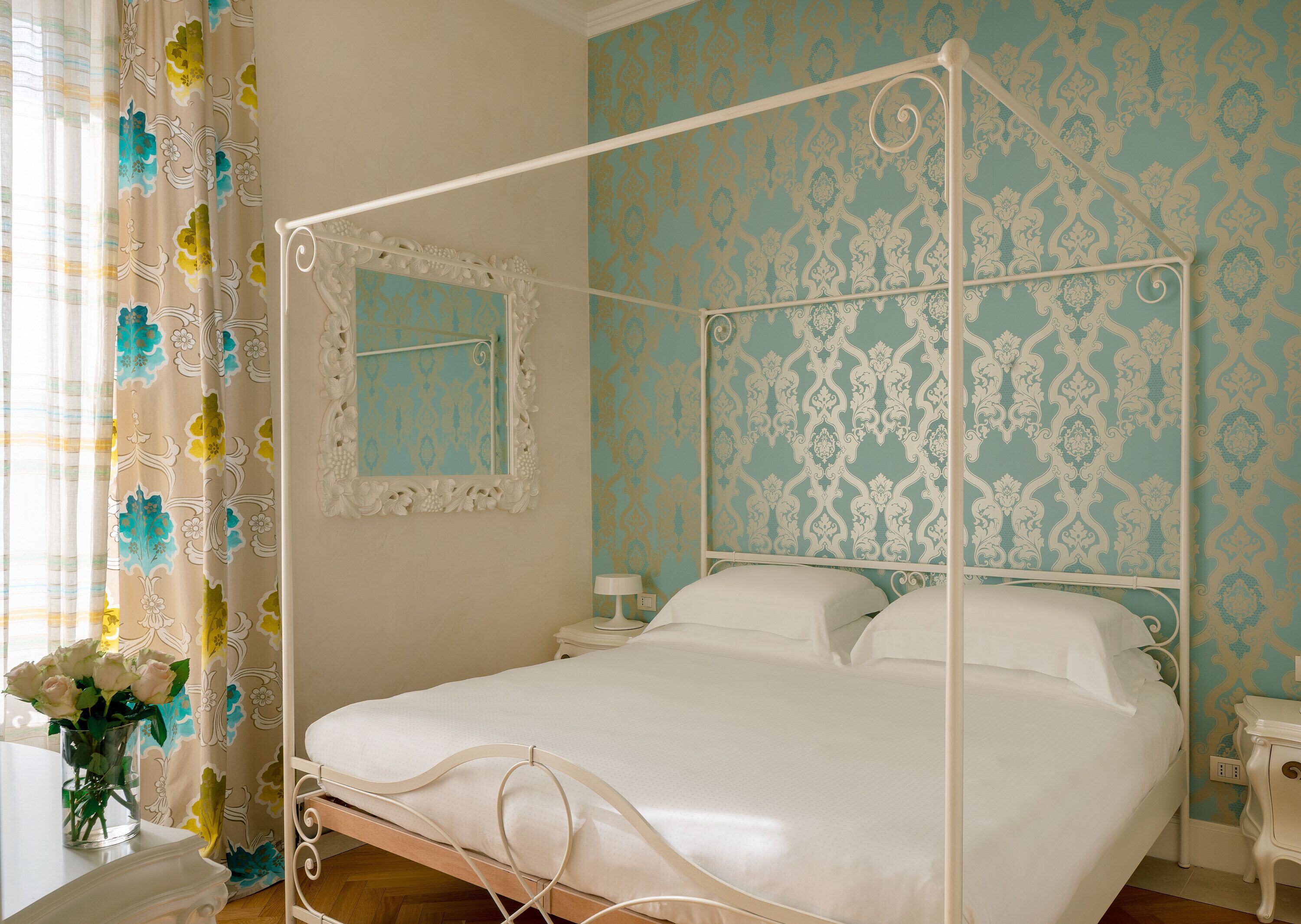 A four-poster bed with white bedding, a mirror above it, and a floral-patterned wall.