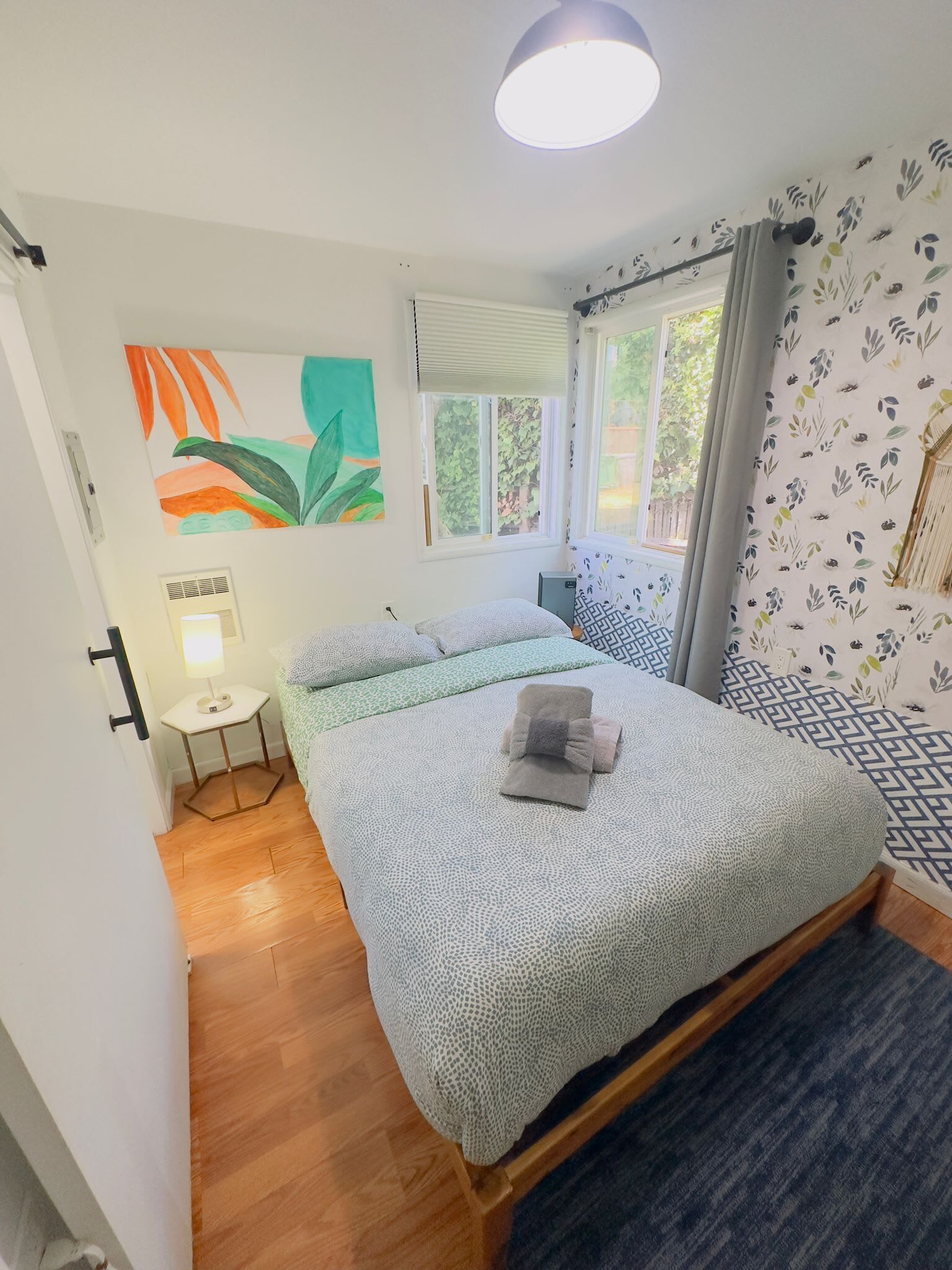 Quarto com cama, mesa de cabeceira, abajur, janela com cortinas e parede com padrão floral.