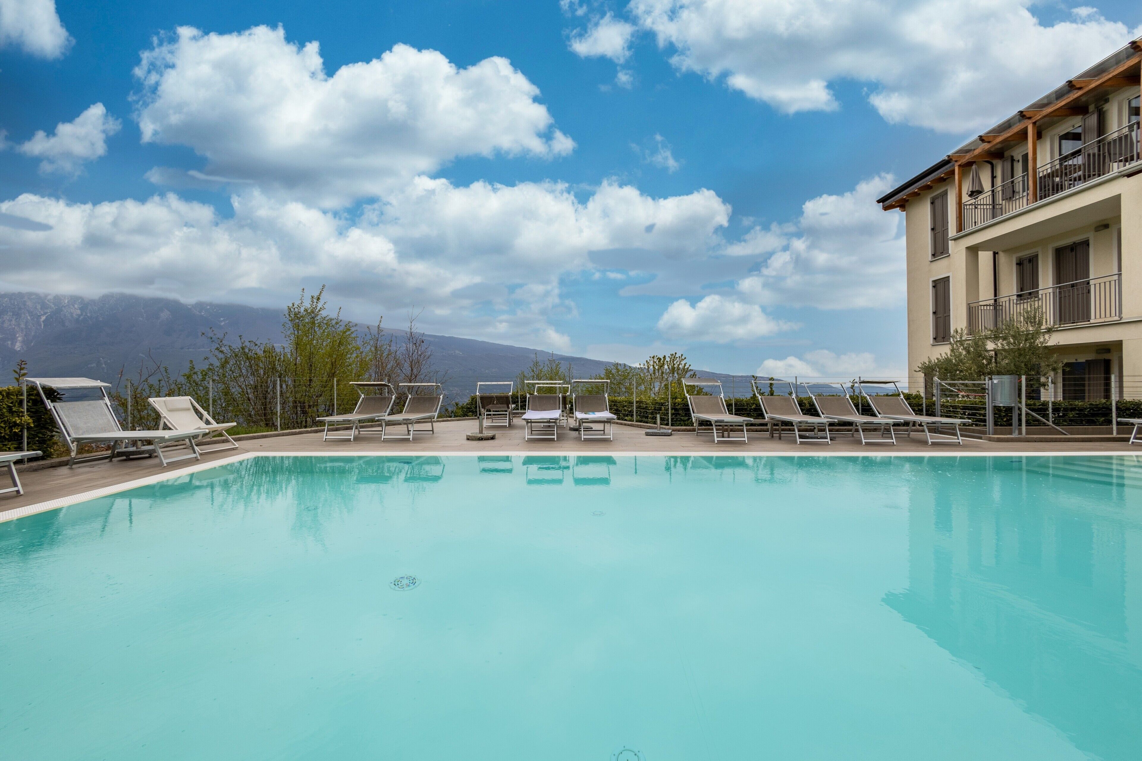 Ein Swimmingpool mit Liegestühlen, ein Gebäude mit Balkonen und Berge im Hintergrund.