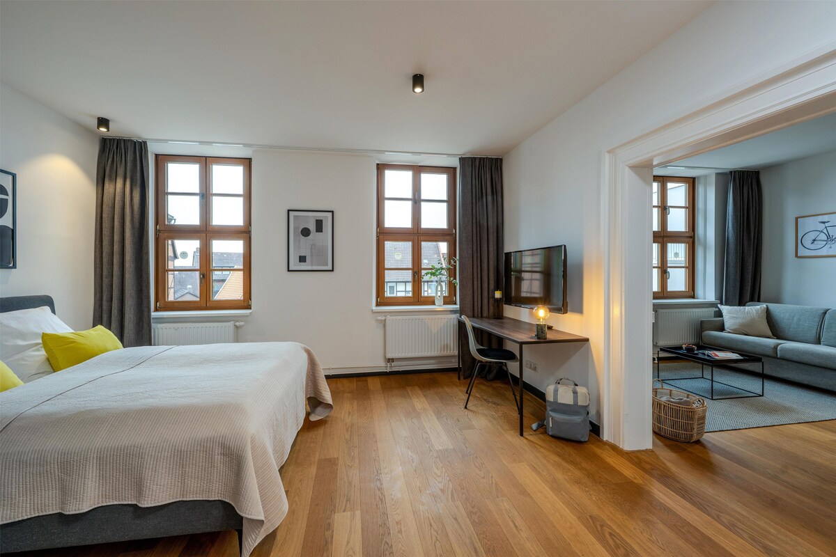A modern bedroom with a bed, desk, and seating area, featuring large windows and a view of buildings.