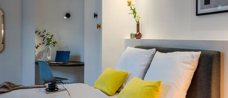 Een moderne slaapkamer met een bed, een bureau met een laptop, een stoel en een wandvaas met bloemen.