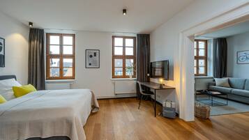 Un dormitorio moderno con cama, escritorio y zona de estar, con ventanas amplias y vista a edificios.