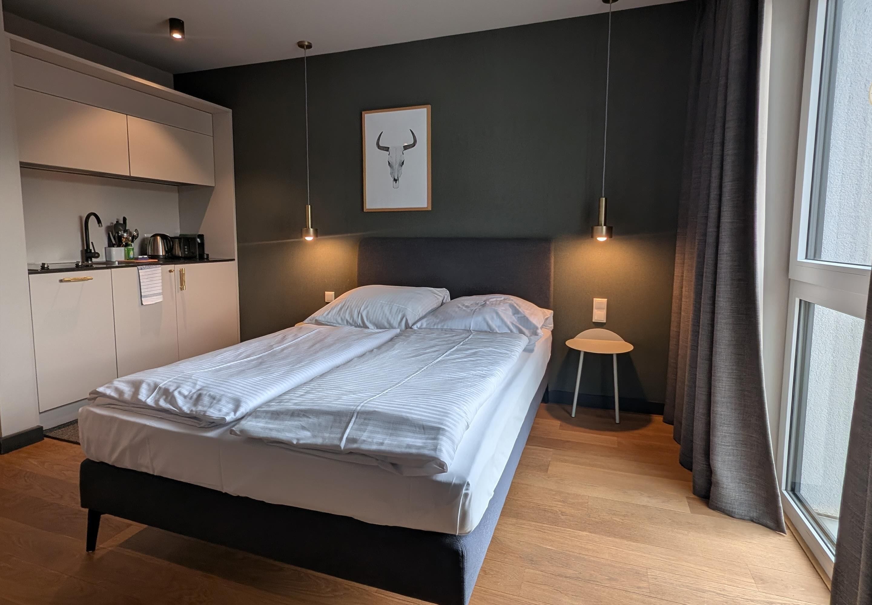 A bedroom with a bed, bedside lamps, a headboard, a window with curtains, a kitchen area with cabinets, and a wall-mounted picture.