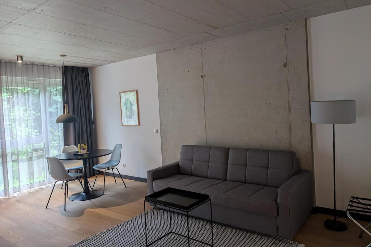 Ett modernt vardagsrum med en grå soffa, ett runt bord med stolar, en golvlampa och ett stort fönster med gardiner.