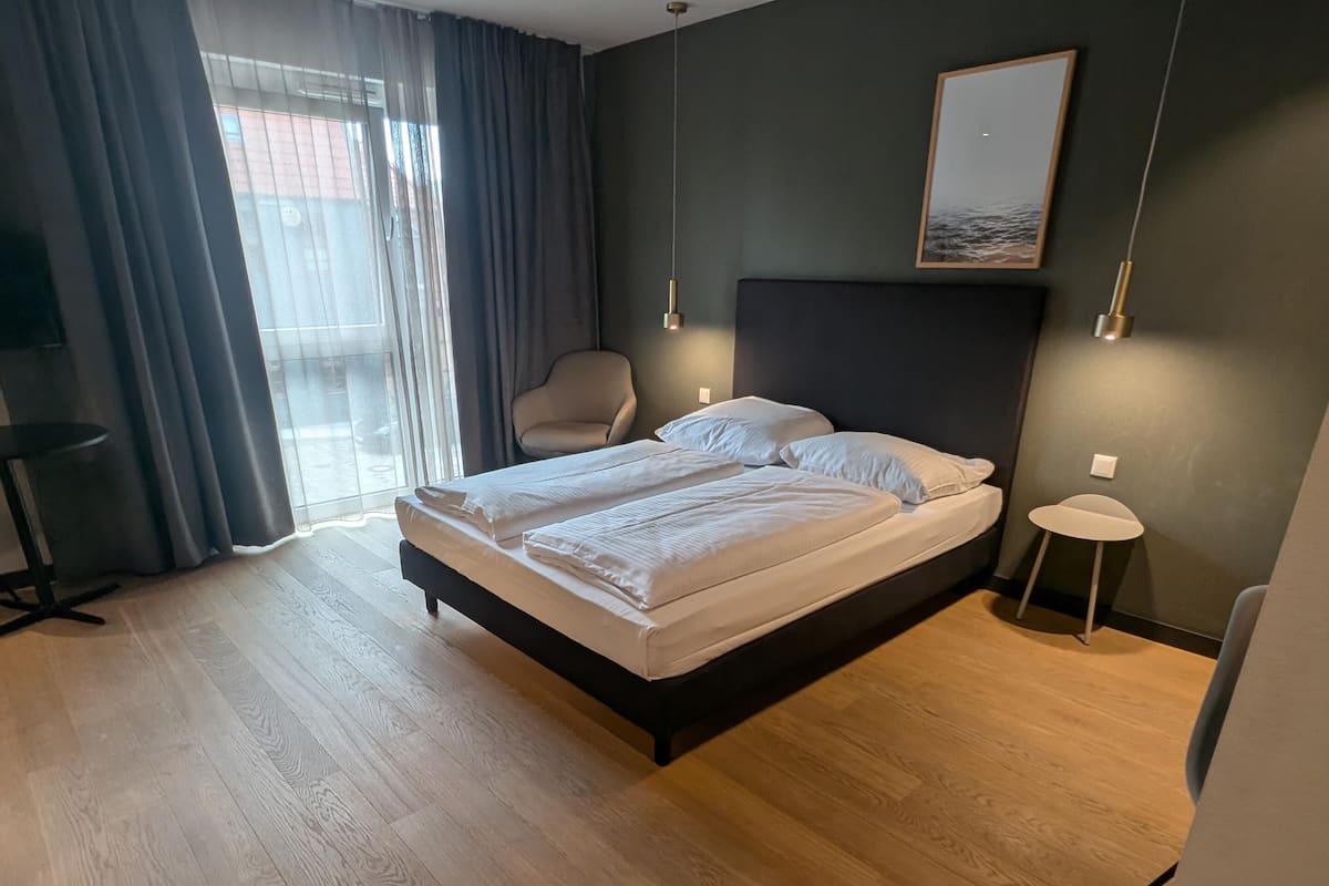 Ett modernt hotellrum med en stor säng, en stol, ett litet bord och en utsikt genom ett fönster med gardiner.