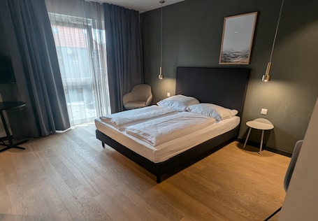 A modern hotel room with a large bed, a chair, a small table, and a view through a window with curtains.. ipartment Wolfsburg @HOME by Best Western
