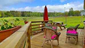 Une terrasse en bois, agrémentée de chaises et d’une table en osier, donnant sur un champ verdoyant bordé d’une clôture et de quelques arbres.