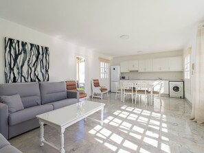 A modern living room with a grey sofa, a white coffee table, and a colorful abstract painting on the wall. There is a kitchen area with white cabinets, a microwave, and a washing machine.