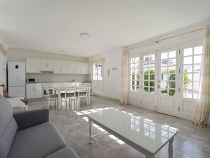 A spacious living area with a dining table and chairs, a sofa, a kitchen with white cabinets, and a large sliding glass door leading to a garden.