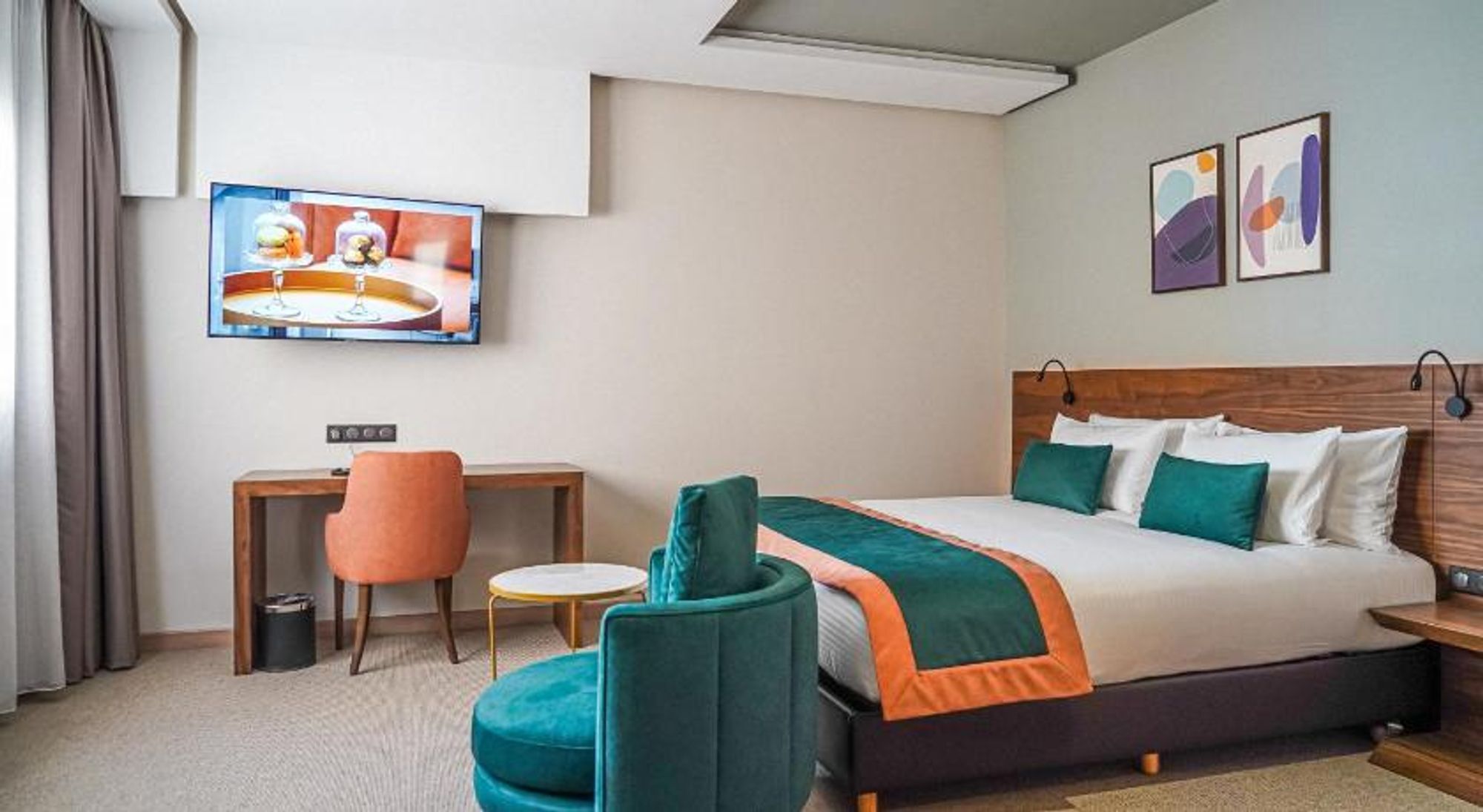 A hotel room with a bed, a desk, a chair, a TV, and artwork on the wall.
