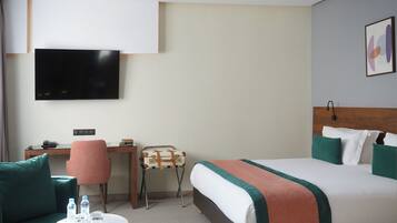 A hotel room with a bed, a desk, a chair, a TV, and a small table with water bottles.