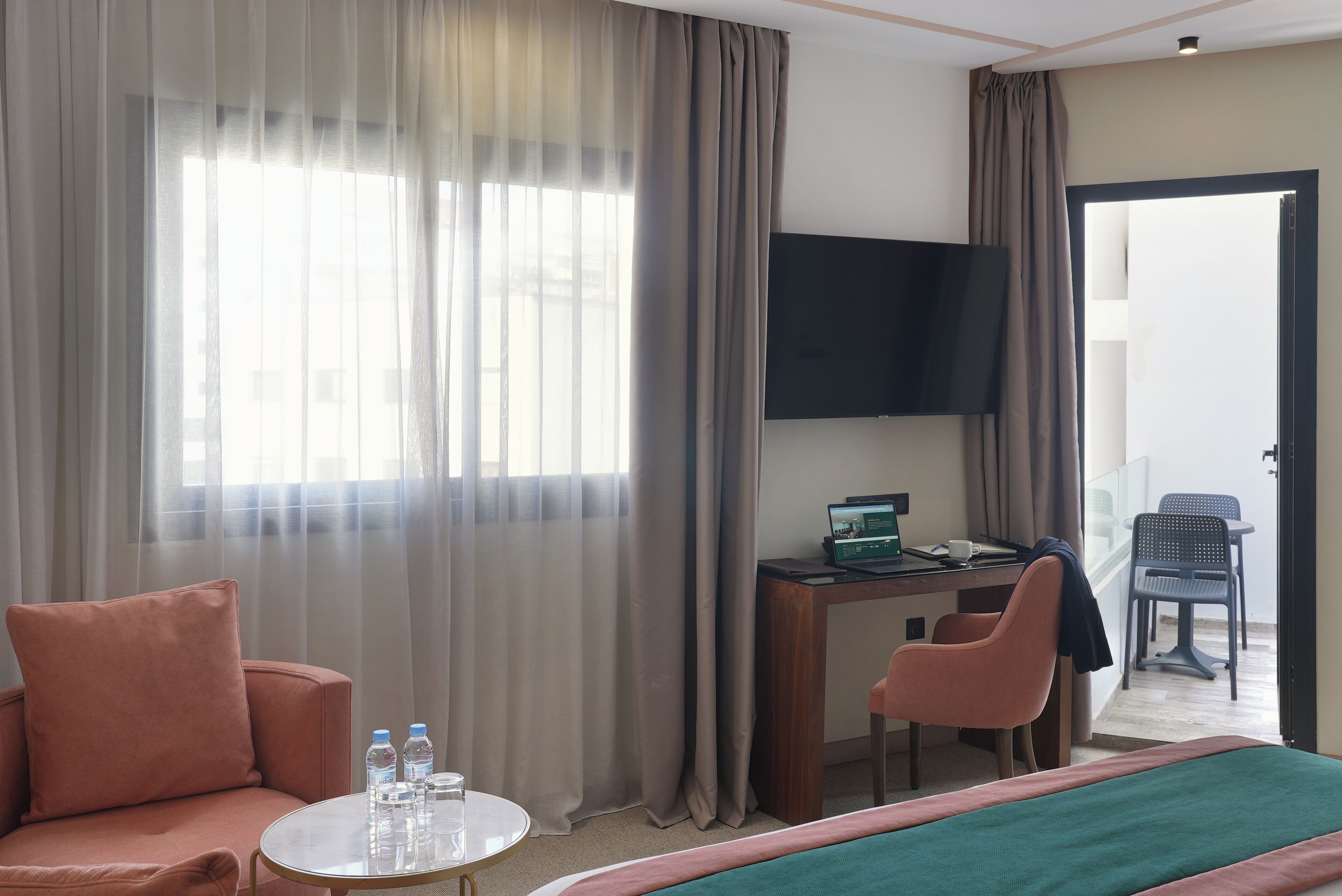A hotel room with a bed, a desk with a laptop, a chair, a sofa, and a TV on the wall.