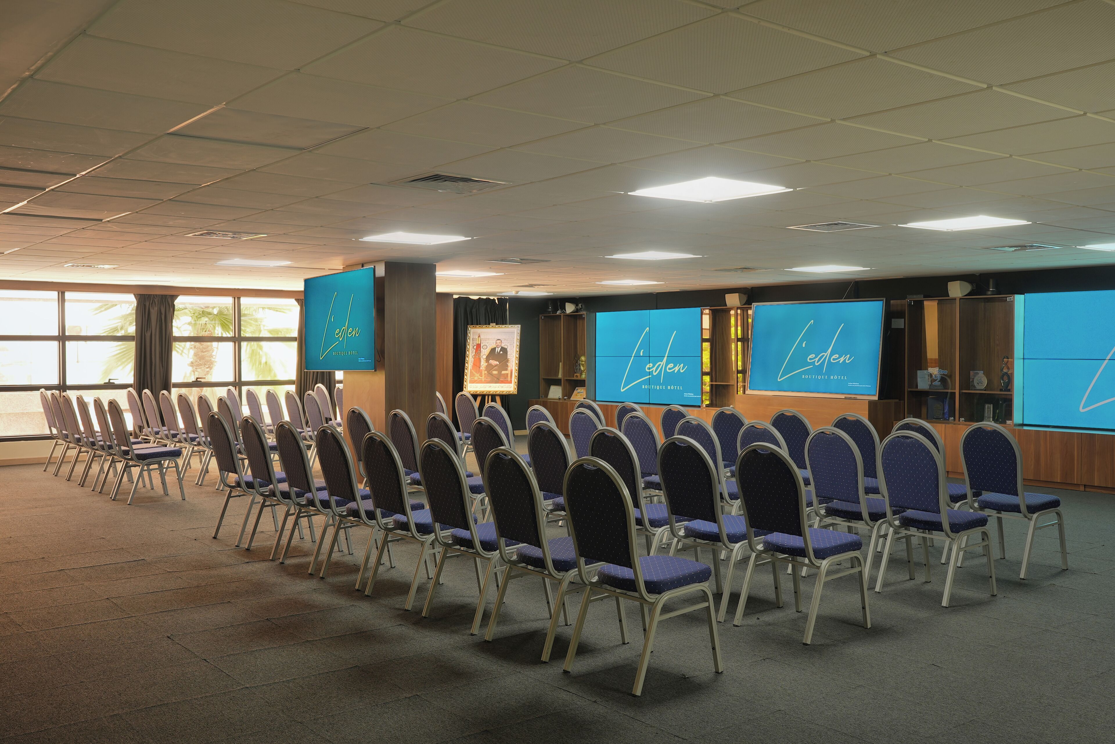 Un salón de conferencias con filas de sillas, ventanas amplias y pantallas mostrando el nombre del hotel.