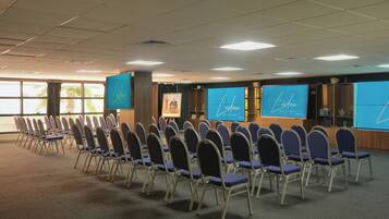 Un salón de conferencias con filas de sillas, ventanas amplias y pantallas mostrando el nombre del hotel.