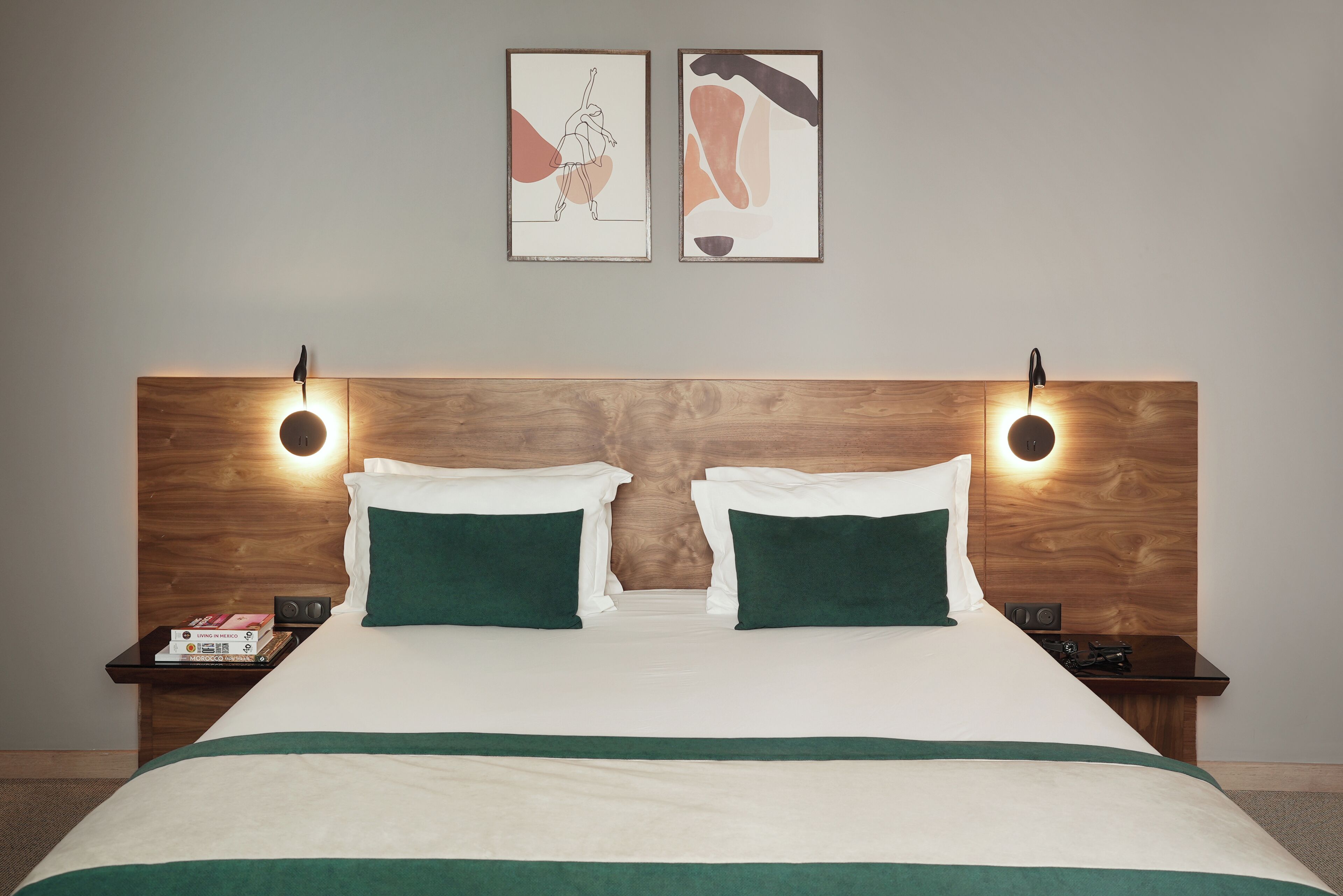Habitación de hotel con cabecera de madera, dos mesitas de noche, cama con ropa de cama verde y blanca, y dos obras de arte enmarcadas en la pared.