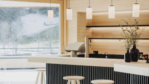 A modern interior with a wooden ceiling, a bar area with stools, and a large window offering a view of a snowy landscape. - Peterhof Chalets (Zwischenwasser)
