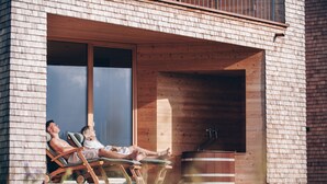 A modern building with a brick exterior, large windows, and a wooden deck with lounge chairs. - Peterhof Chalets (Zwischenwasser)
