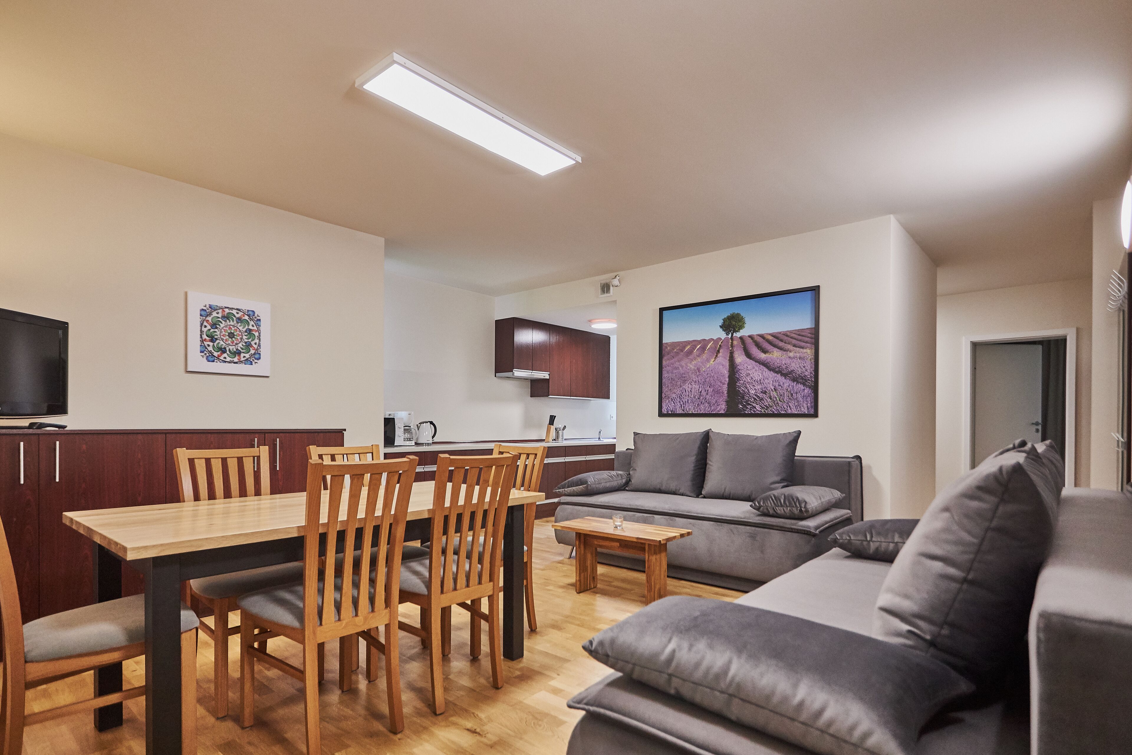 Ein modernes Wohnzimmer mit grauem Sofa, Holzesstisch und Stühlen. Im Hintergrund ist eine Küchenzeile mit Schränken und einem Flachbildfernseher zu sehen.