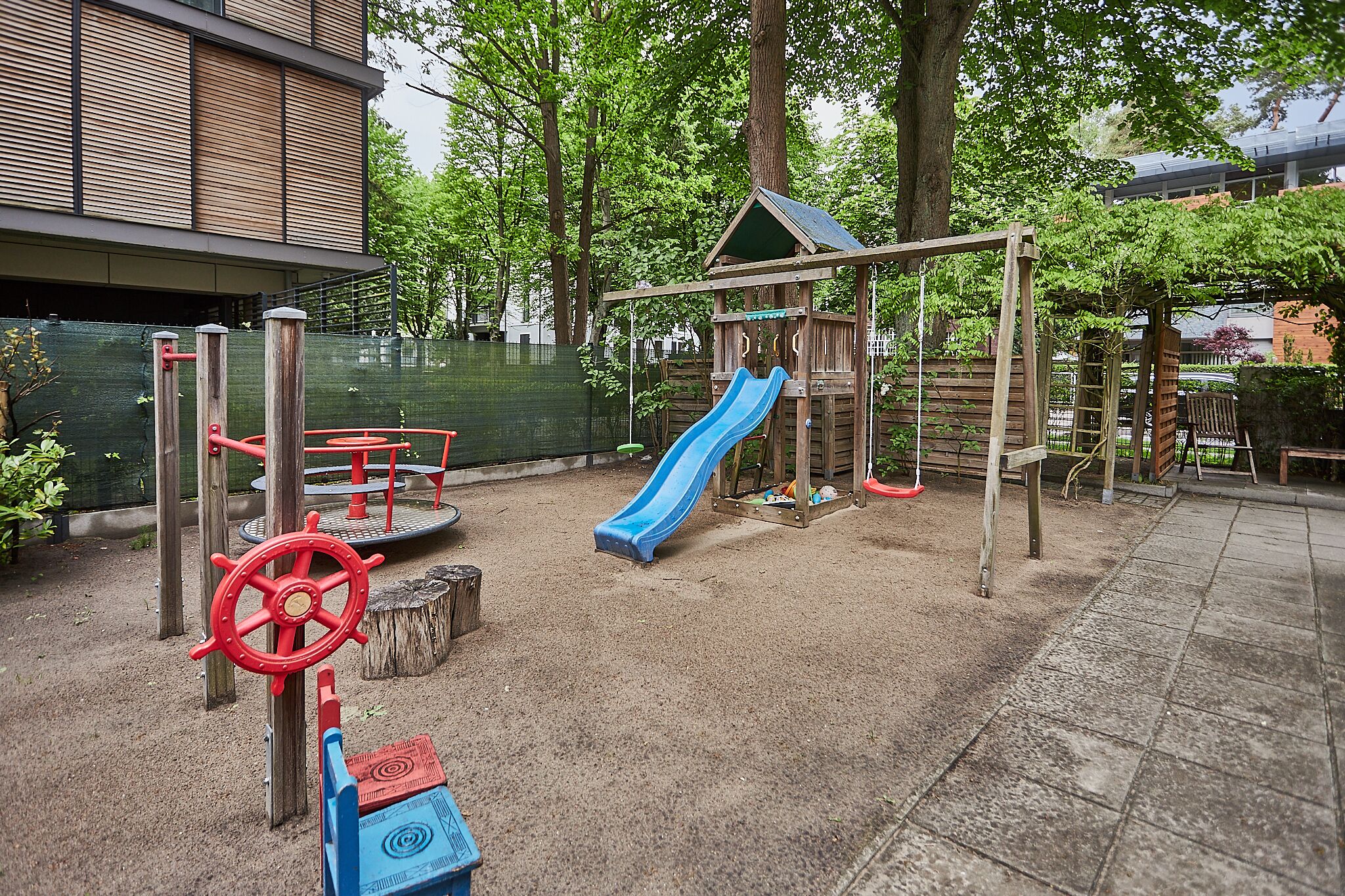 A playground with a slide and climbing structures, surrounded by trees and a building in the background.