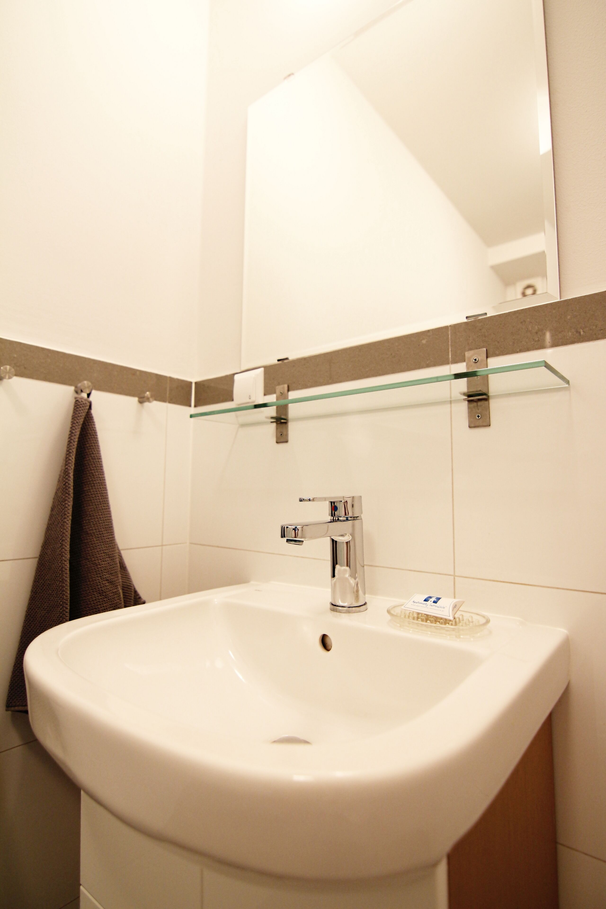 Un baño con un lavabo blanco, un estante de vidrio encima y una toalla colgada en la pared.