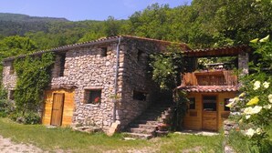 A stone house with a wooden balcony, surrounded by lush greenery.