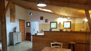 A rustic kitchen with wooden beams, a wooden table with chairs, a refrigerator, and a window with a view.