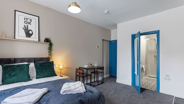 A hotel room with a bed, a desk, a chair, a small table with a plant, a framed picture on the wall, and a bathroom door.