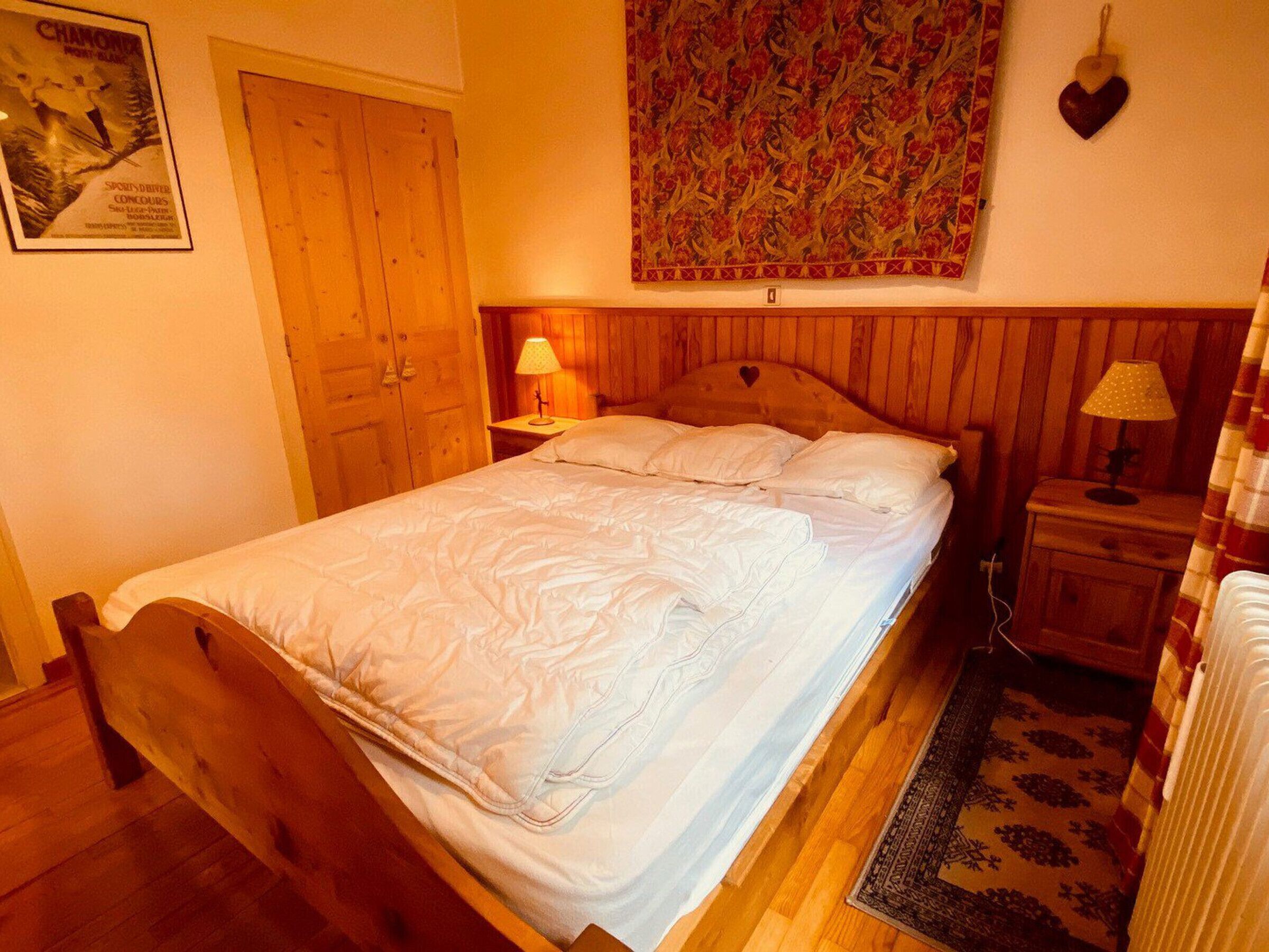 A bedroom with a wooden bed, a nightstand with a lamp, a decorative wall hanging, and a poster on the wall.