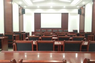 Un salón de conferencias con filas de sillas de madera, una mesa grande al frente y una pantalla de proyección.