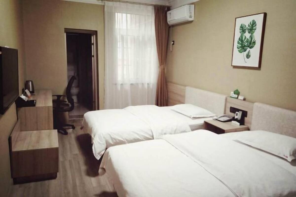 Une chambre d’hôtel avec deux lits, un bureau, une chaise, un appareil de climatisation et une plante décorative accrochée au mur.