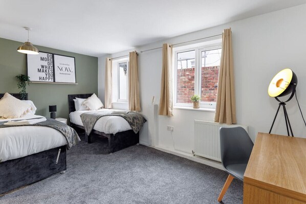 Une chambre avec deux lits, un bureau, une chaise, une lampe et une fenêtre avec des rideaux.