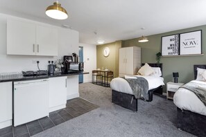 Une chambre d’hôtel compacte comprenant un coin cuisine, un lit avec une tête de lit, une table de chevet et une horloge murale.