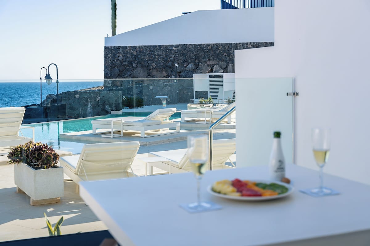 Un área de piscina en la azotea con sillones de descanso, una mesa de comedor con una fuente de frutas y dos copas de vino, y vistas al océano.