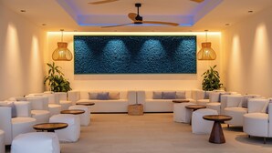 Uma área de estar moderna com sofás, pufes e mesas redondas, com destaque para uma grande obra de arte texturizada na parede e iluminação ambiente.