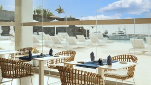 Uma área de refeições com cadeiras e mesas de vime, com vista para uma piscina e espreguiçadeiras.