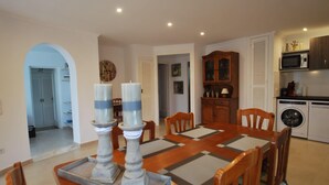A dining area with a wooden table, chairs, and a chandelier. There is a kitchen area with appliances and cabinets in the background.