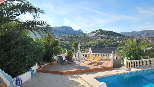 A poolside area with a view of mountains, a white fence, and a patio with chairs and umbrellas.