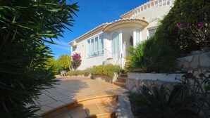 A house with a stone pathway leading to a white door, surrounded by lush greenery and flowering plants.