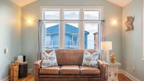 A living room with a brown leather sofa, a glass side table, and a view of blue houses through large windows. - Fresh and Salty (Carolina Beach)