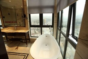 A modern bathroom with a large bathtub, a marble vanity, and a large window offering a city view.