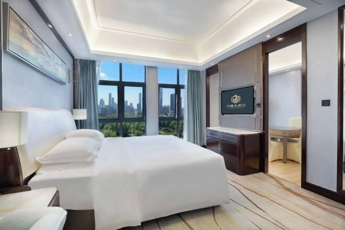 A modern hotel room with a large bed, a TV, a desk, and a view of the cityscape.