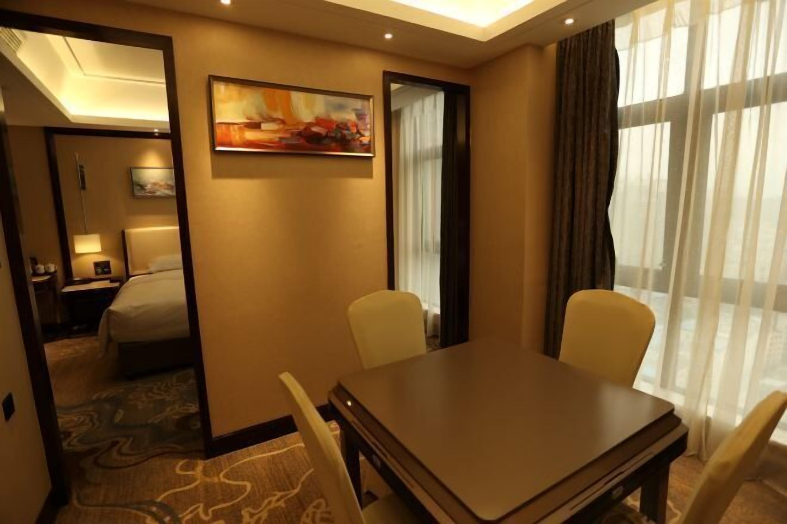 A hotel room with a bed, a desk, and a chair. There is a painting on the wall and a window with curtains.