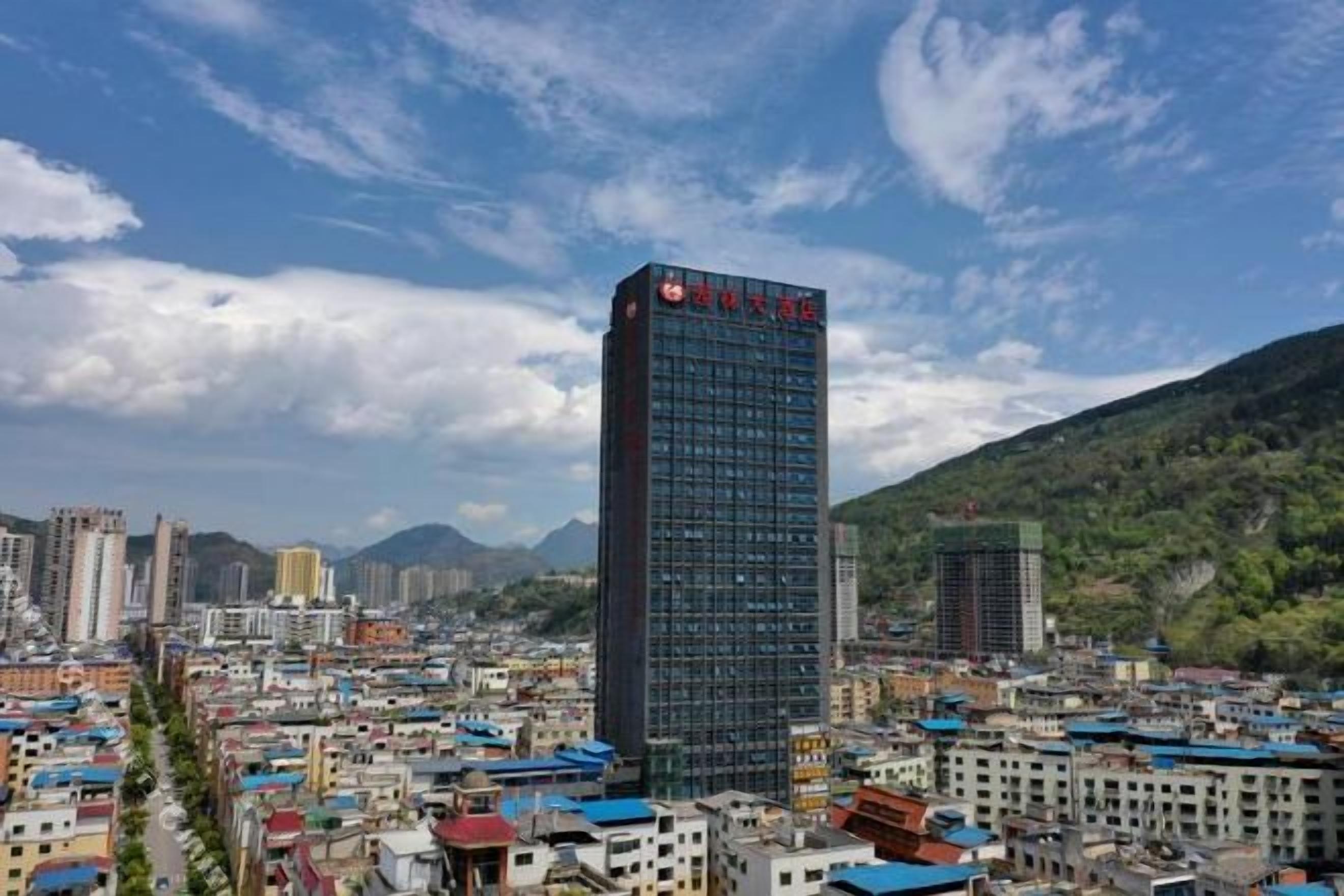 Un paisaje urbano con un edificio moderno destacado, que tiene un letrero rojo, rodeado de edificios más pequeños y un telón de fondo montañoso.