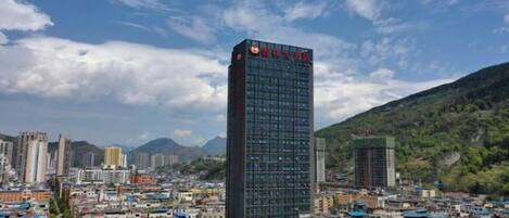Un paisaje urbano con un edificio moderno destacado, que tiene un letrero rojo, rodeado de edificios más pequeños y un telón de fondo montañoso.
