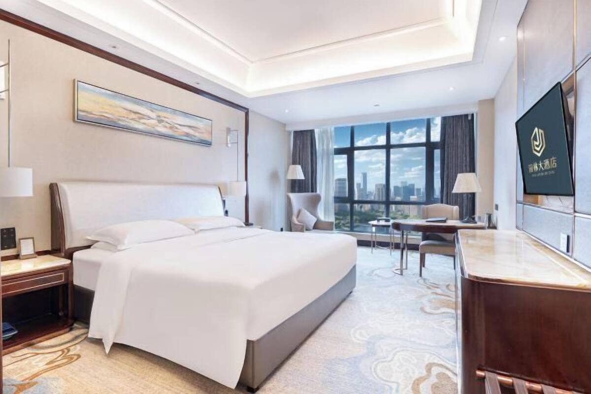 A hotel room with a large bed, a desk, a chair, a TV, and a view of the city.