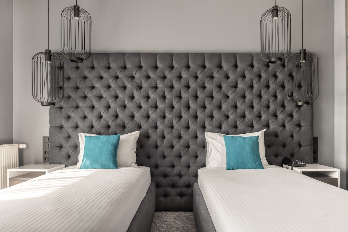 Un dormitorio moderno con cabecera capitonada, dos camas con ropa blanca, almohadas turquesa y lámparas colgantes negras.