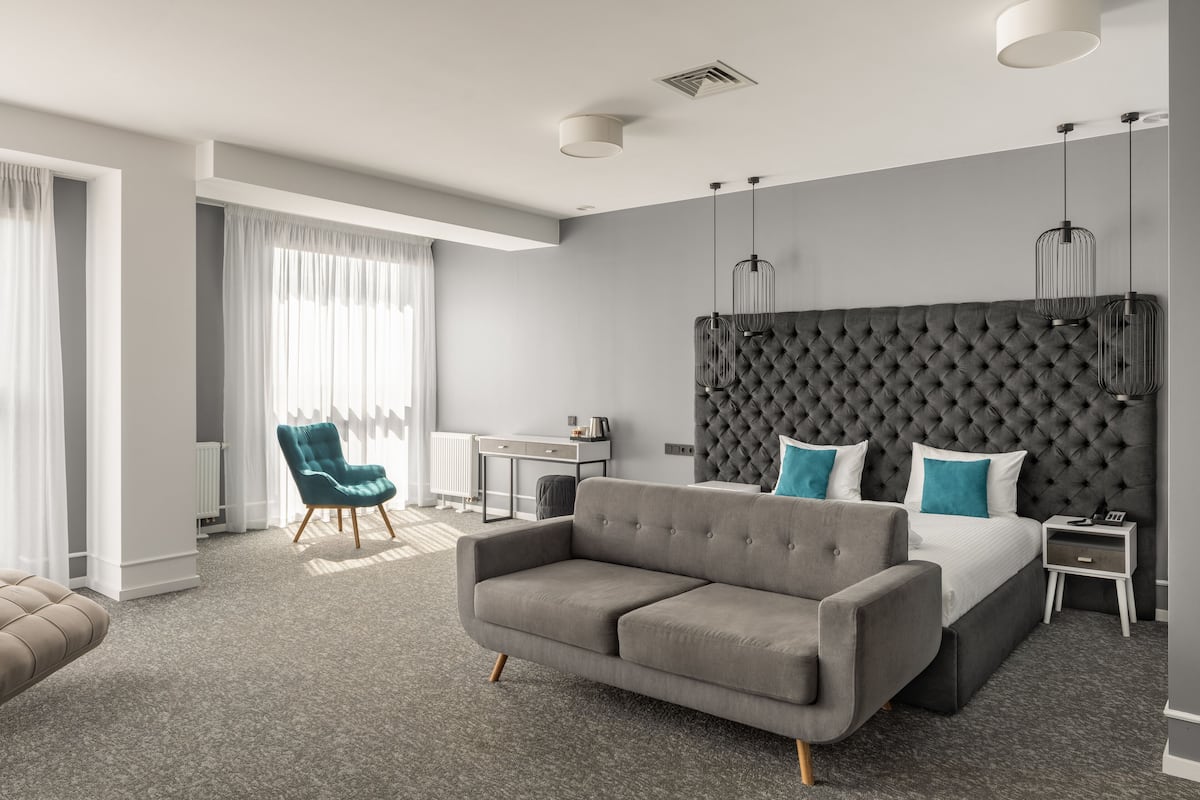 Una habitación de hotel moderna con un cabecero capitonado, un sofá gris, un sillón verde azulado, un escritorio pequeño y una mesita de noche con una lámpara.